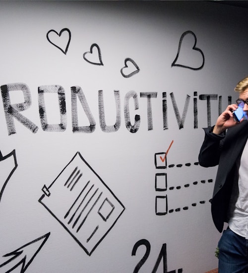A person dressed in business attire is talking on a mobile phone while standing next to a wall decorated with motivational drawings and the word 'Productivity' in bold letters. The wall features symbols like hearts, arrows, and checklists.