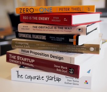 A stack of business and entrepreneurship books on a table. The titles are oriented towards strategies, innovation, and startup guidance, including 'Zero to One', 'Ego is the Enemy', and 'The Startup Owner's Manual'. The setting appears to be in a modern office or study environment.