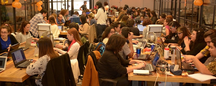 A large group of people are gathered in a spacious, well-lit office or coworking space. They are seated at long wooden tables working on laptops. Some are collaborating in small groups while others are focused on their individual tasks. The atmosphere appears to be busy and productive with a friendly and collaborative vibe. There are decorations like orange balloons and string lights that add to the lively environment.