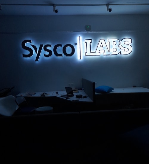 A dimly lit room features a prominent illuminated sign displaying the words 'Sysco LABS.' Below the sign are office desks with various electronic devices, including laptops and cables. The lighting creates a cool and professional atmosphere.